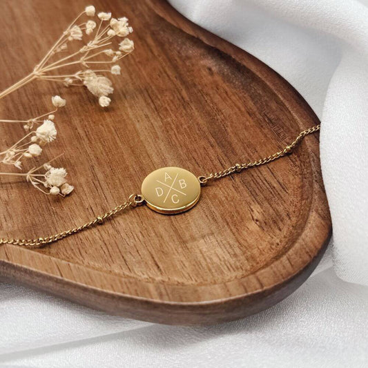 Goldenes Armband mit vier gravierten Initialen auf einem Holzbrett