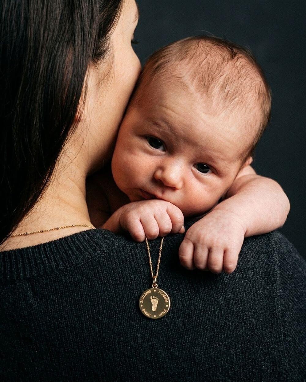 Baby liegt bei Mama über der Schulter und hält eine goldene Mizodia Fußabdruck mit Geburtsdatenkette